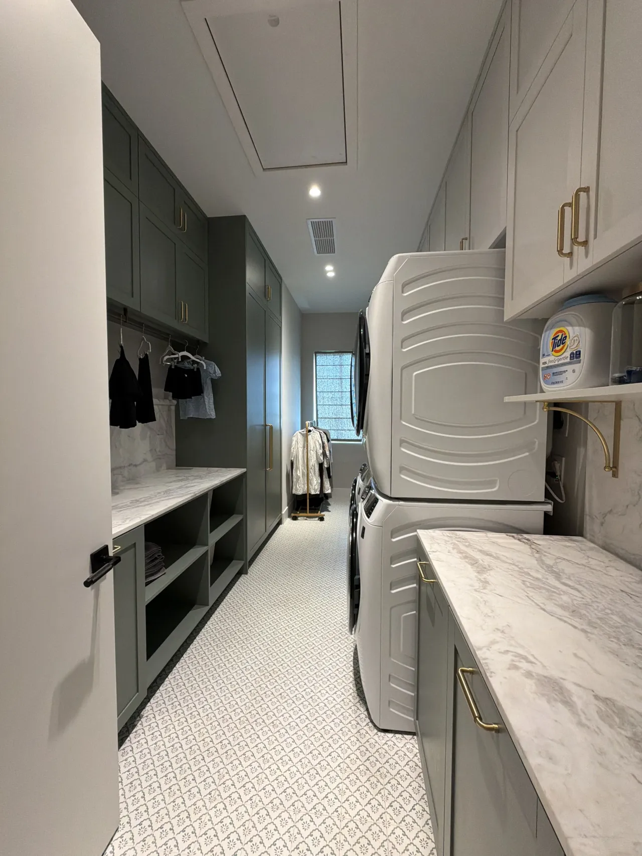Narrow laundry room seen down its length, green cabinets with open cubbies at left, white uppers, stone counters and a stacked washer/dryer at right
