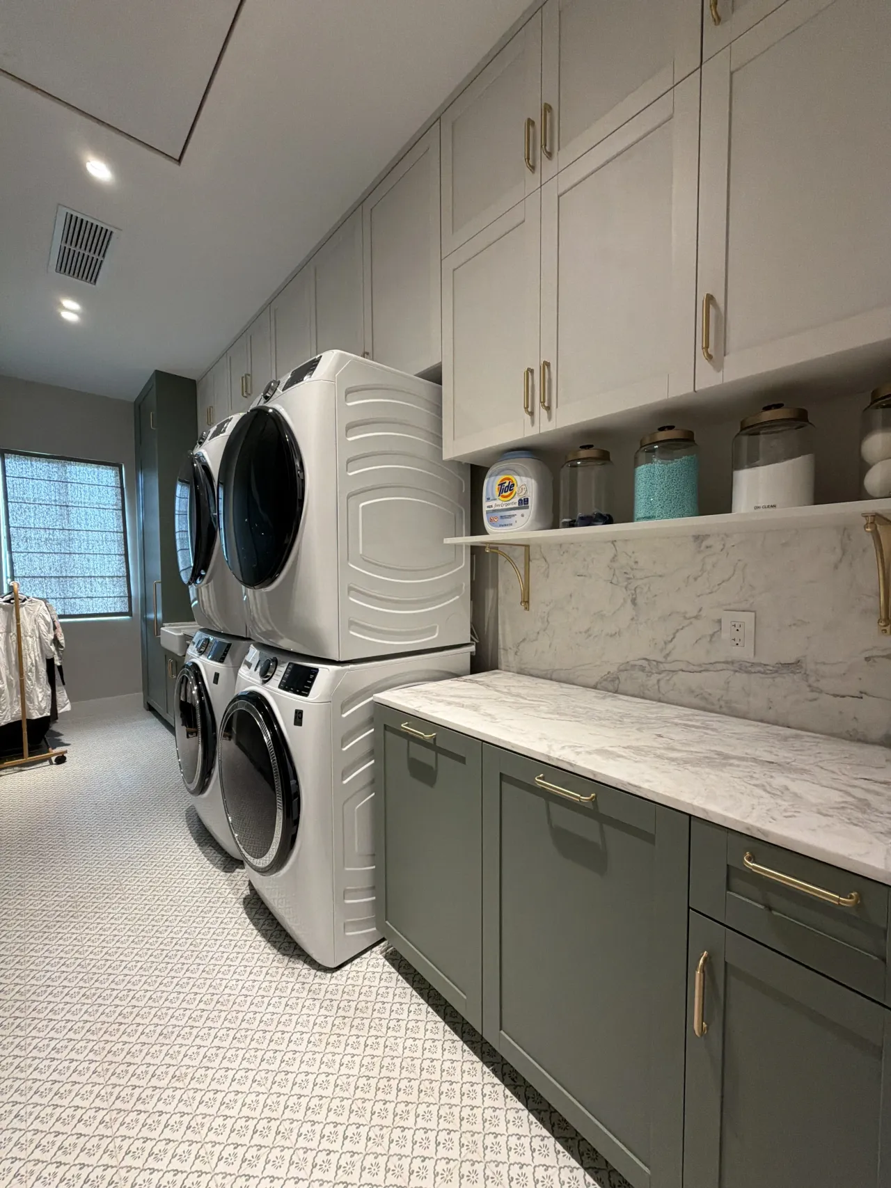 Laundry room with sage green melamine shaker-style base cabinets, gold-tone hardware, veined stone countertop and backsplash, stacked white washer/dryer