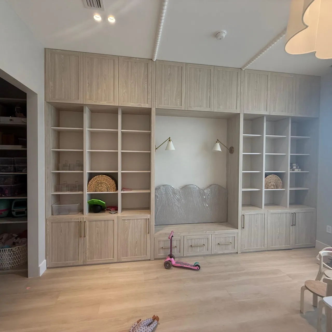 Light wood built-in shelving wall with open cubbies, a reading nook bench, gold-tone swing-arm sconces, and lower drawers