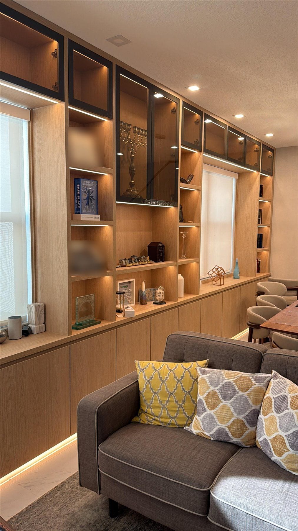 Floor-to-ceiling light wood-grain built-in with lit open shelves, black-framed glass display cabinets, and flush lower doors with toe-kick lighting