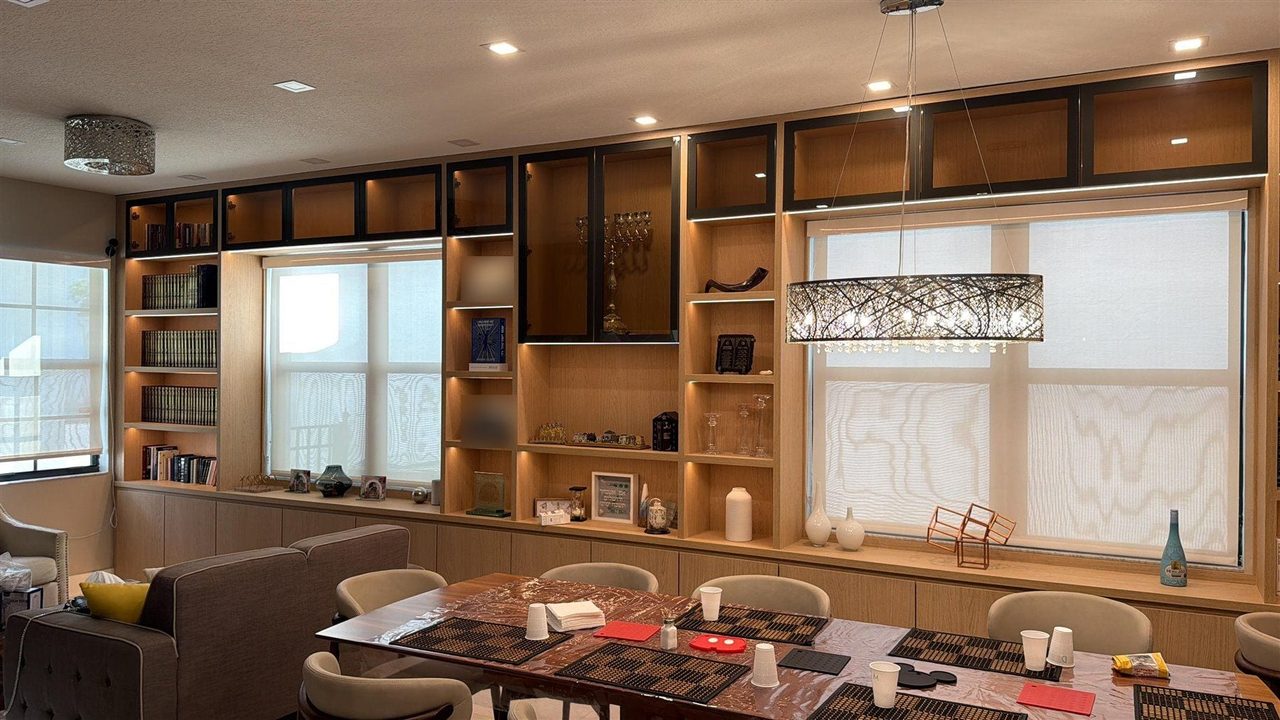 Long light wood-grain built-in wall with lit open shelving, black-framed glass upper cabinets, and flush lower cabinets running beneath two windows