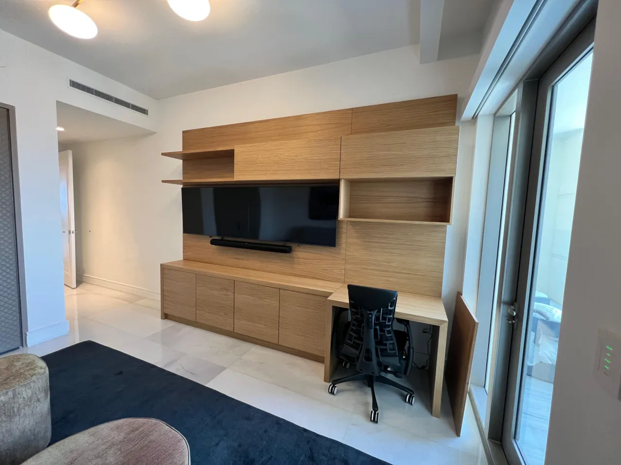 Light wood-grain built-in wall unit with open shelves, wall-mounted TV and soundbar, cabinets below, and a desk return with a task chair to the right