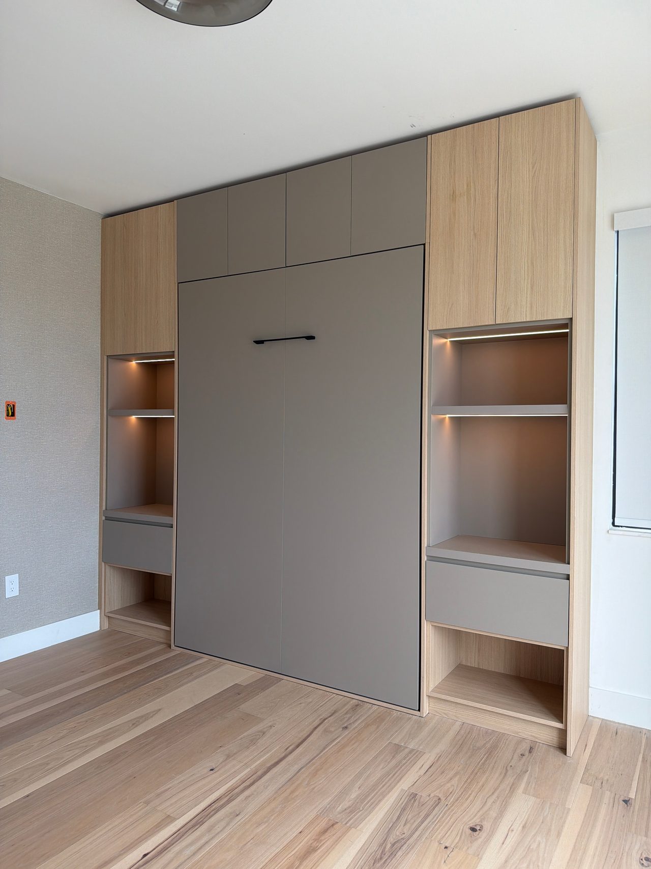Light wood-grain and gray built-in wall unit with a gray fold-down panel, lit open shelves and drawers, textured wallcovering on the wall at left