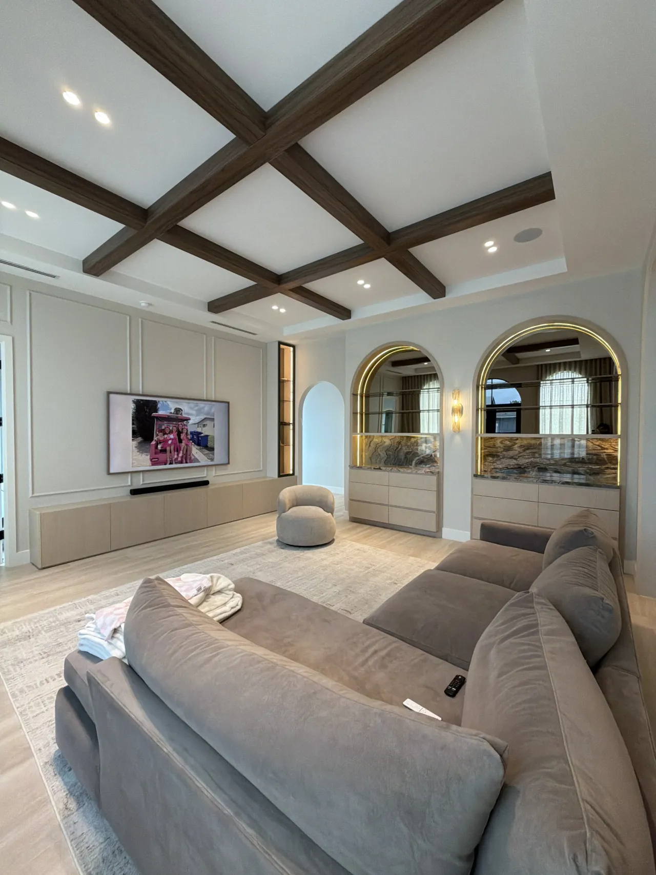 Living room with a crossed dark melamine-clad beam ceiling, paneled TV wall over a light floating credenza, gray sofa, and two arched lit display niches