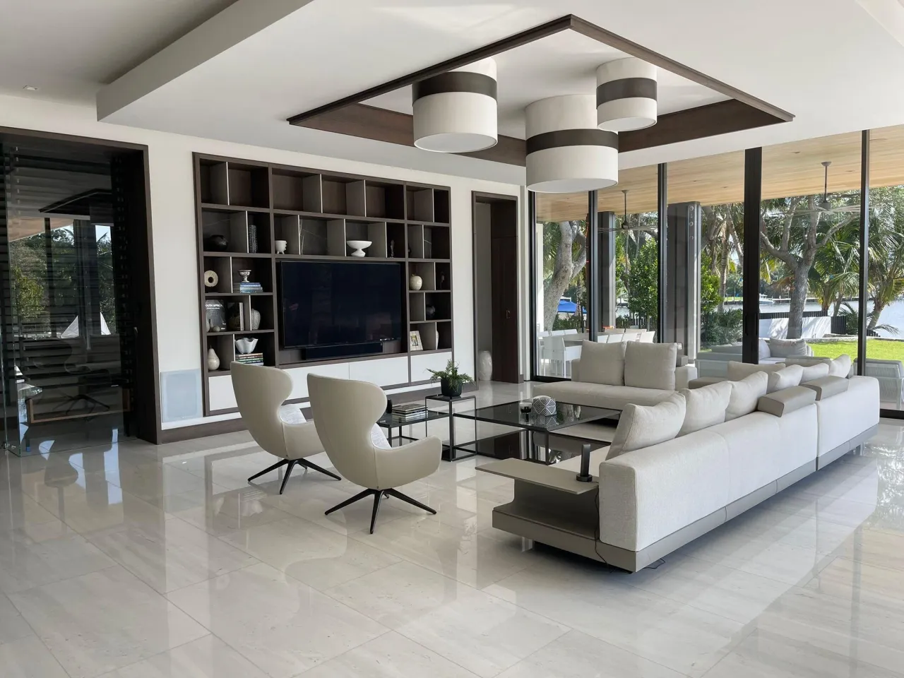 Living room with a floor-to-ceiling open grid shelving unit framing a wall-mounted TV, sectional sofa, drum pendants and polished tile floor