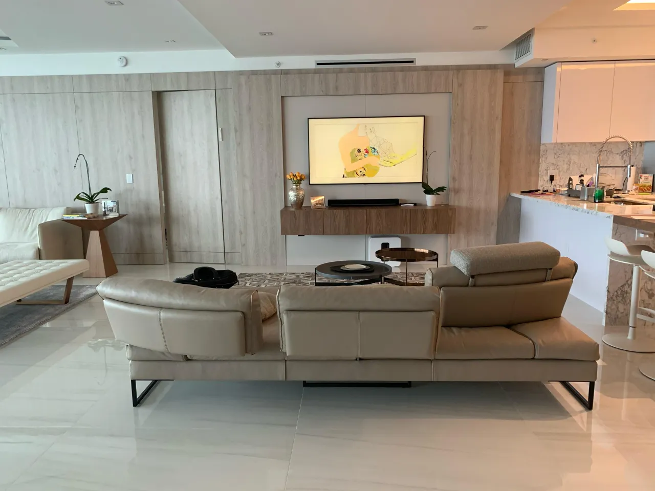 Living room with light wood accent wall behind a wall-mounted TV, framed floating media console, and beige leather-look sectional sofa with view to kitchen