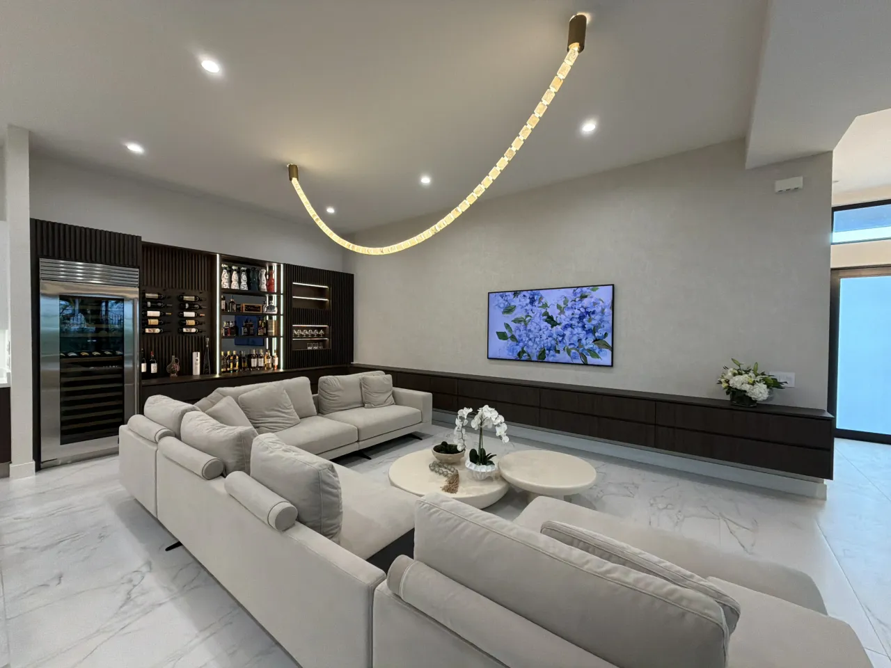 Living room with a large off-white sectional, curved gold-tone tube light, TV over a long dark floating credenza, and a fluted dark bar wall