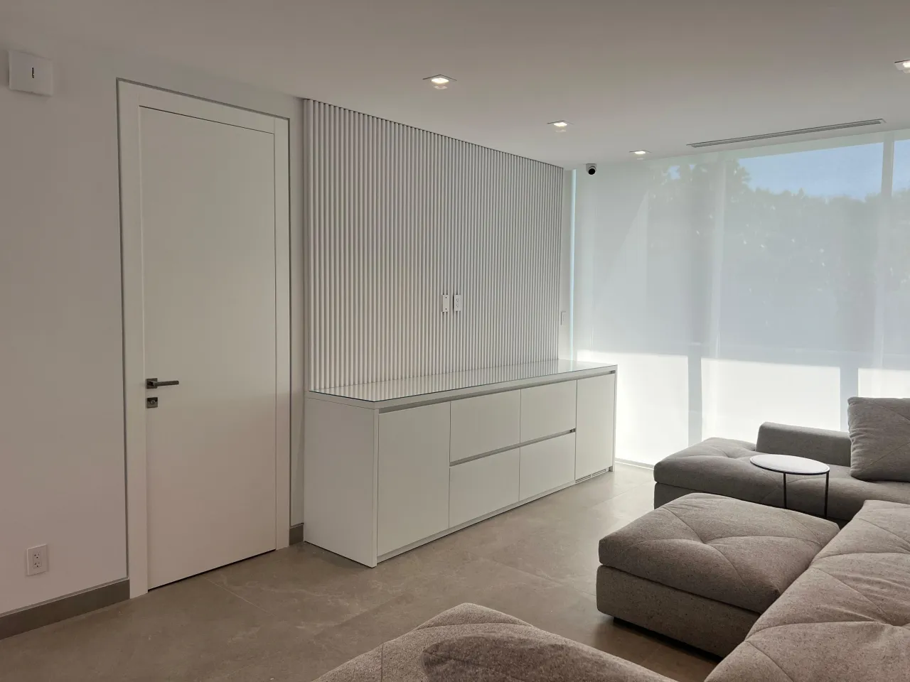 Living room with vertical white slat wall behind a floating white cabinet with drawers, gray sectional sofa in front, and a solid white door beside it