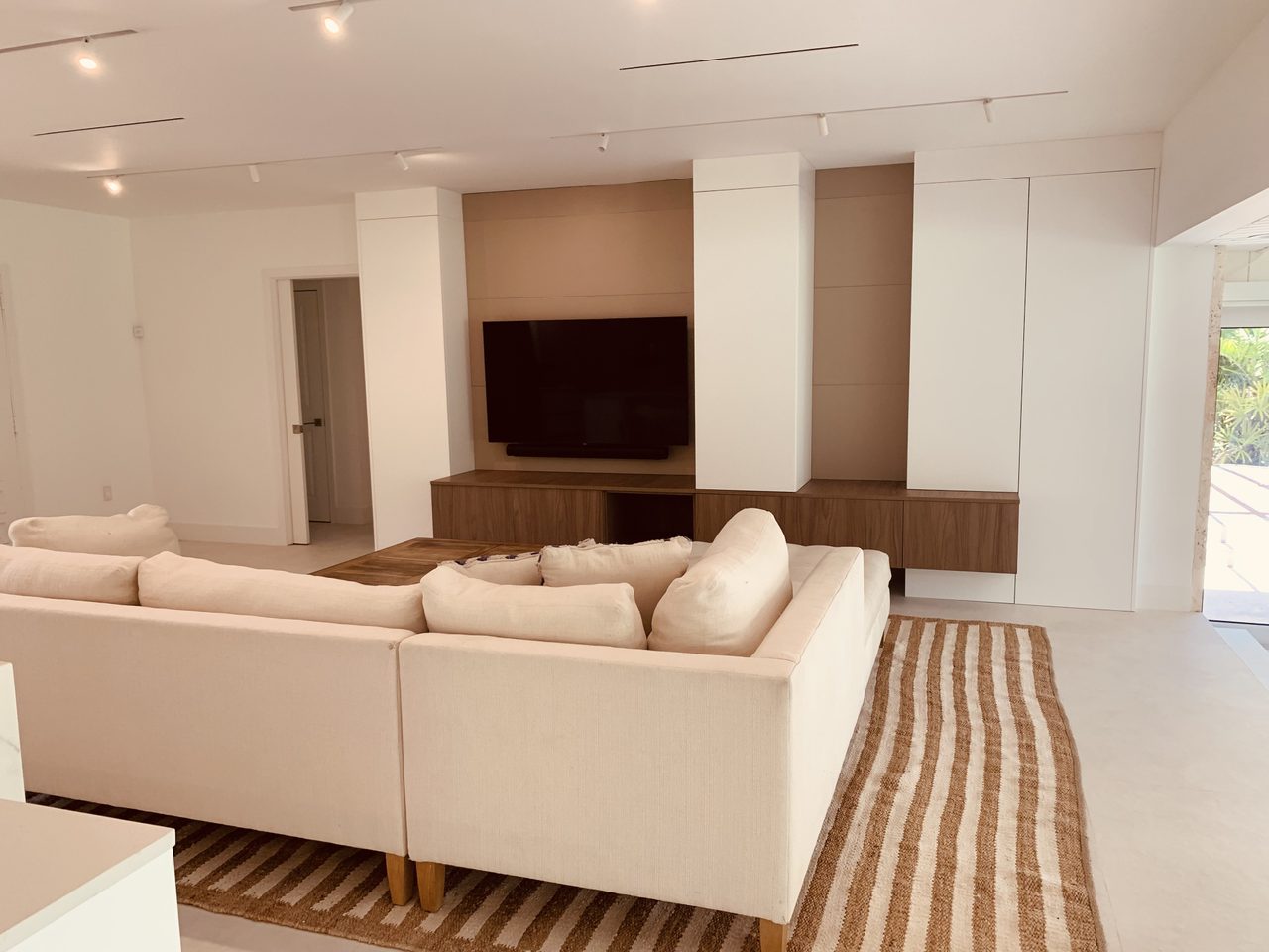 Living room with white floor-to-ceiling cabinetry, recessed TV panel, floating wood-grain console below, cream sectional on a striped rug