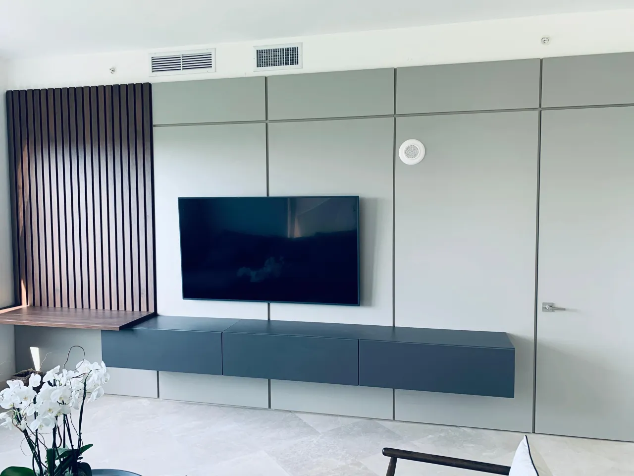 Media wall of pale grey-green flush panels with wall-mounted TV and long dark blue-grey floating console, dark slat panel and wood-grain shelf at left