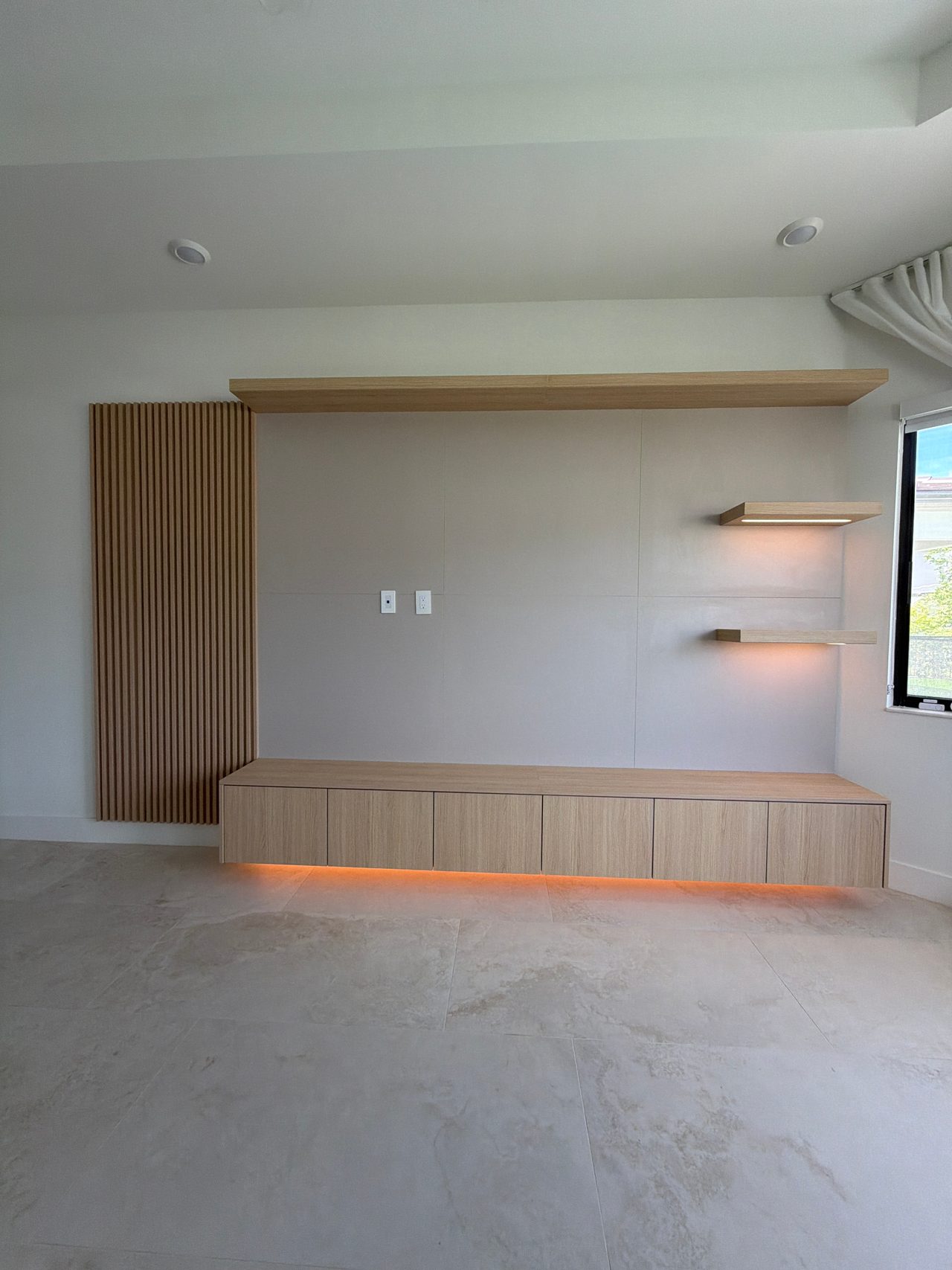 Media wall with a long low wood-grain cabinet lit from beneath in orange, two lit floating shelves, slat panel at left, deep wood-grain header