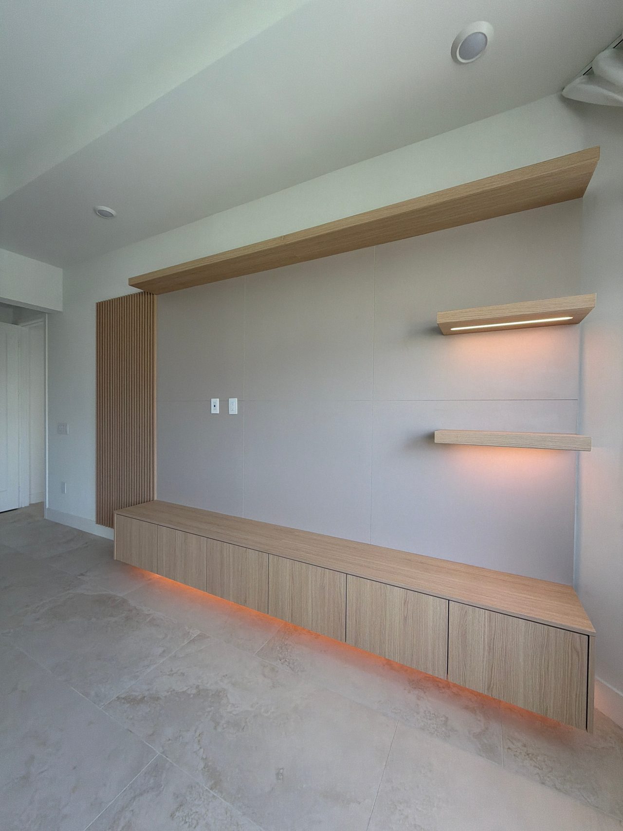Media wall seen at an angle, low wood-grain cabinet with orange underlighting, two lit floating shelves, wood-grain header and slat panel at left
