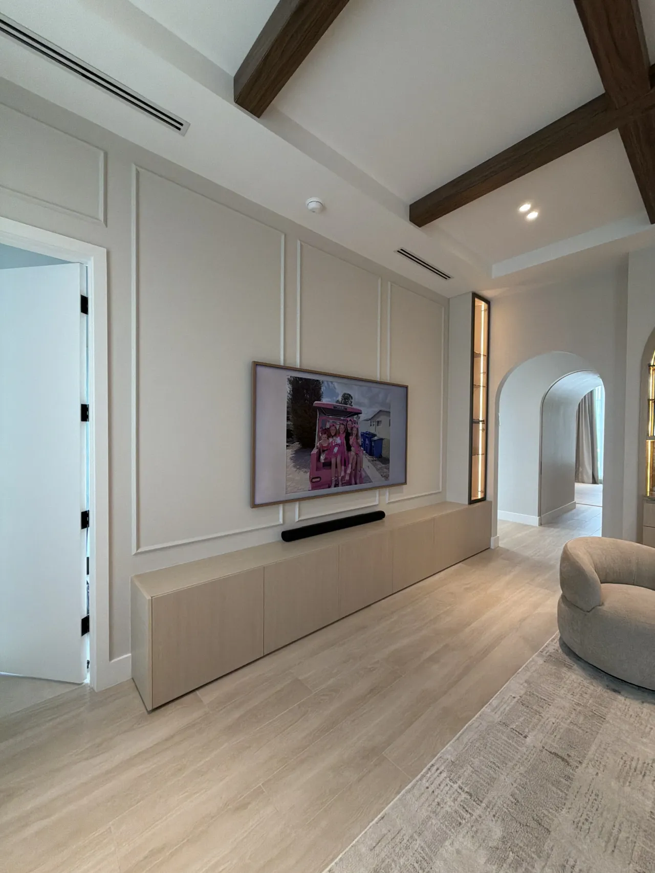 Paneled cream media wall with a wall-mounted TV, soundbar and a long light wood-grain floating credenza, dark melamine-clad ceiling beams above