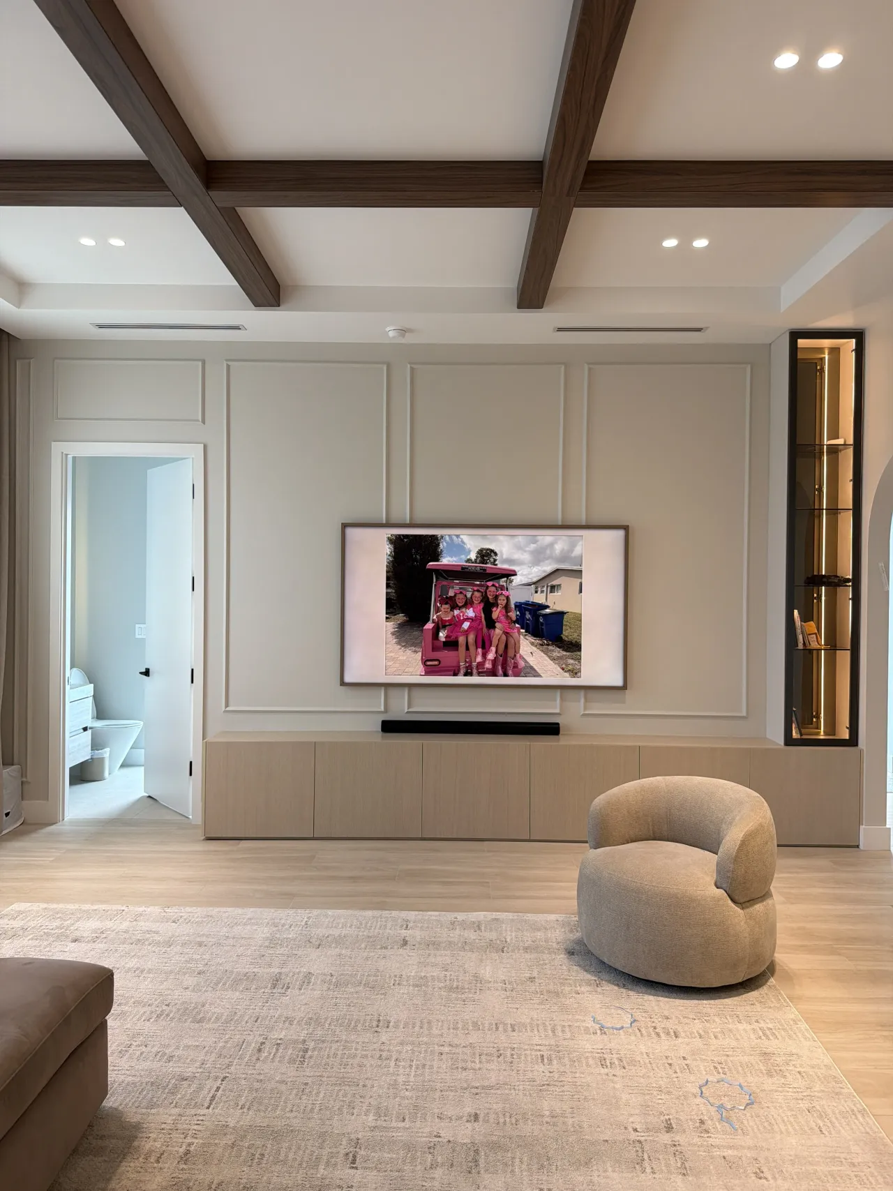 Room with a paneled cream TV wall, mounted TV showing a photo, dark melamine-clad ceiling beams, lit glass display column and a light wood-grain console