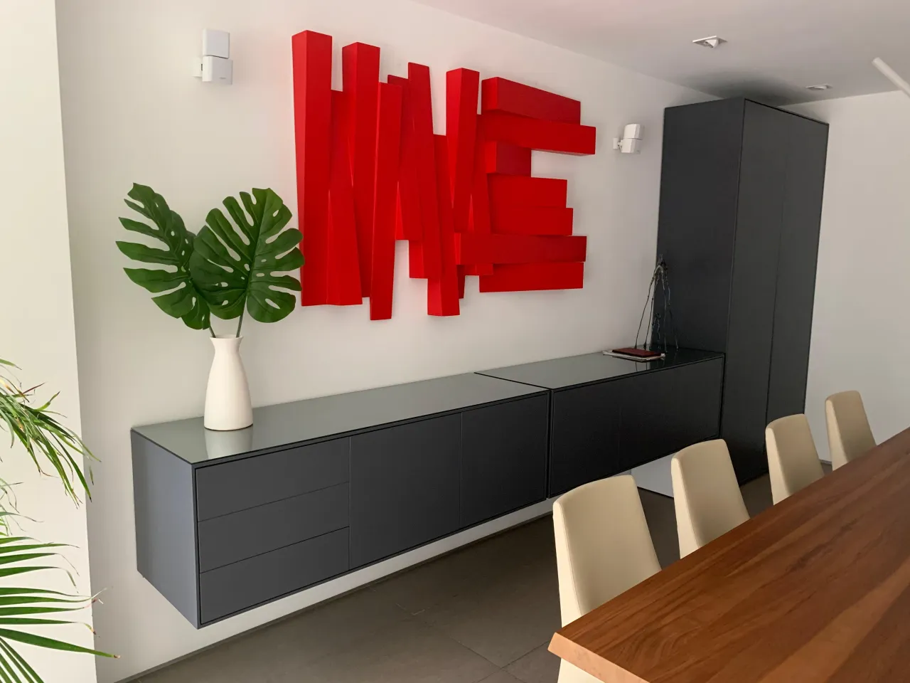 Red 3D lettered wall sculpture above a dark gray floating credenza with drawers, matching tall cabinet, wood-grain table with cream chairs