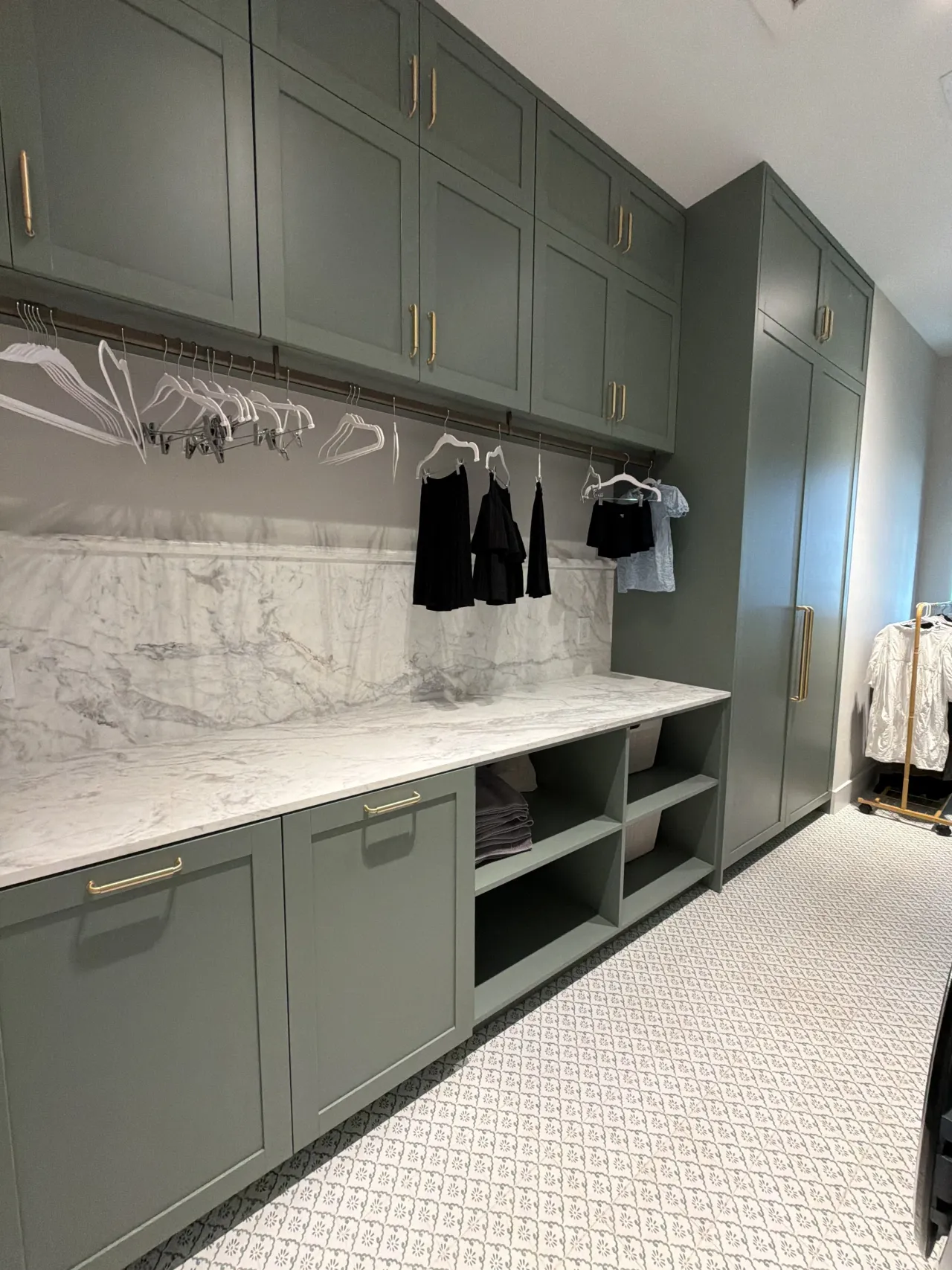 Sage green melamine built-in laundry cabinets with a hanging rod for clothes and open cubby shelving, veined stone backsplash
