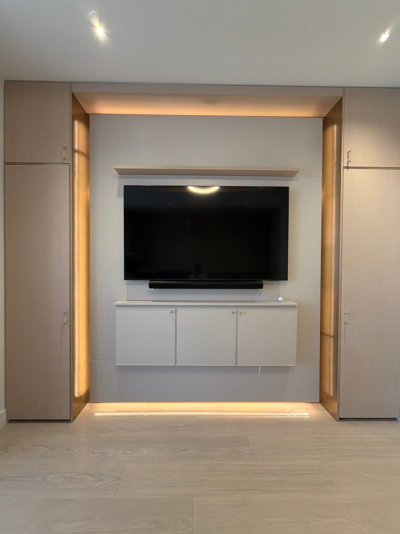 Symmetrical TV wall niche with backlit perimeter trim, wood-grain melamine tall cabinets flanking it and a white wall-hung cabinet under the screen
