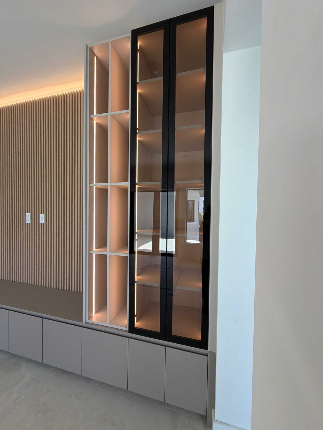 Tall black-framed glass display cabinet with LED-lit shelves beside a white grid shelving column, set above a run of low gray cabinet fronts