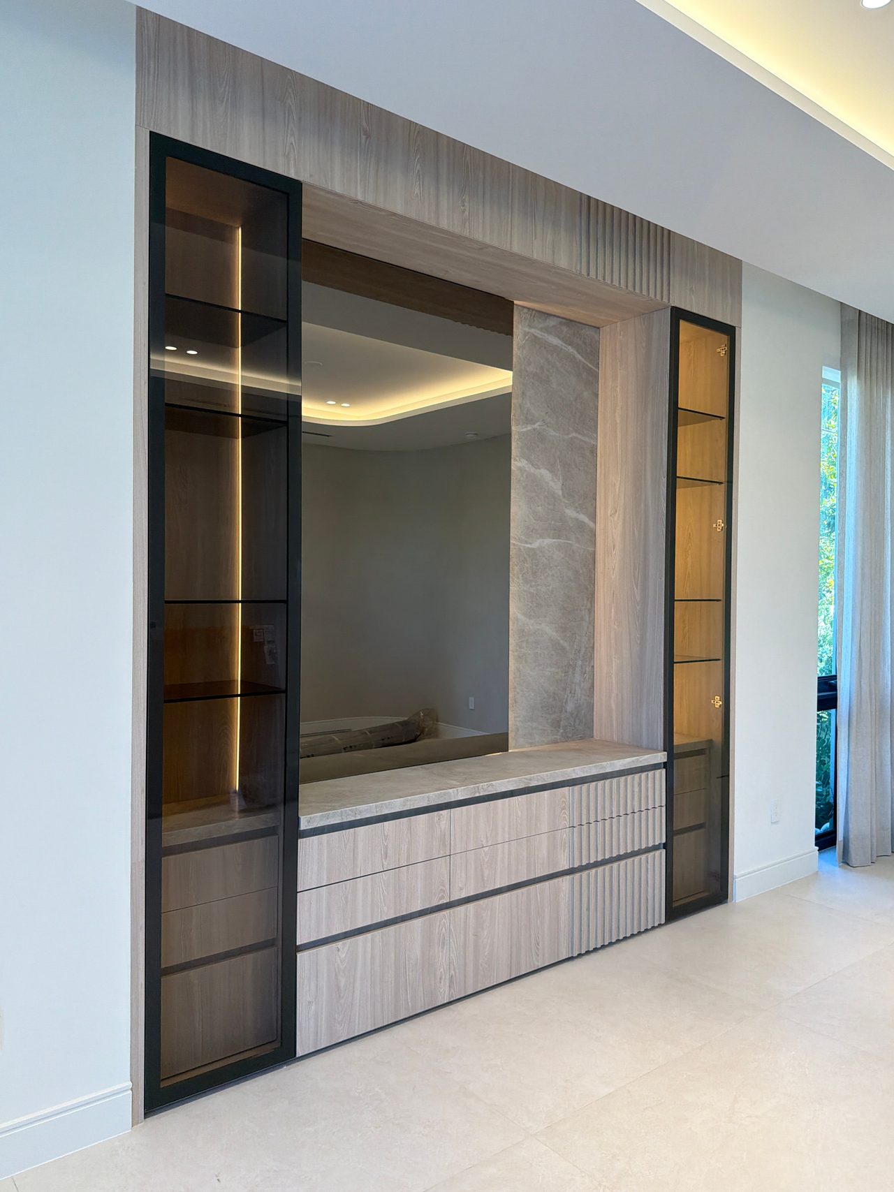 Two tall black-framed glass display cabinets flanking a wood-grain framed pass-through opening, against a veined stone wall, with drawers below