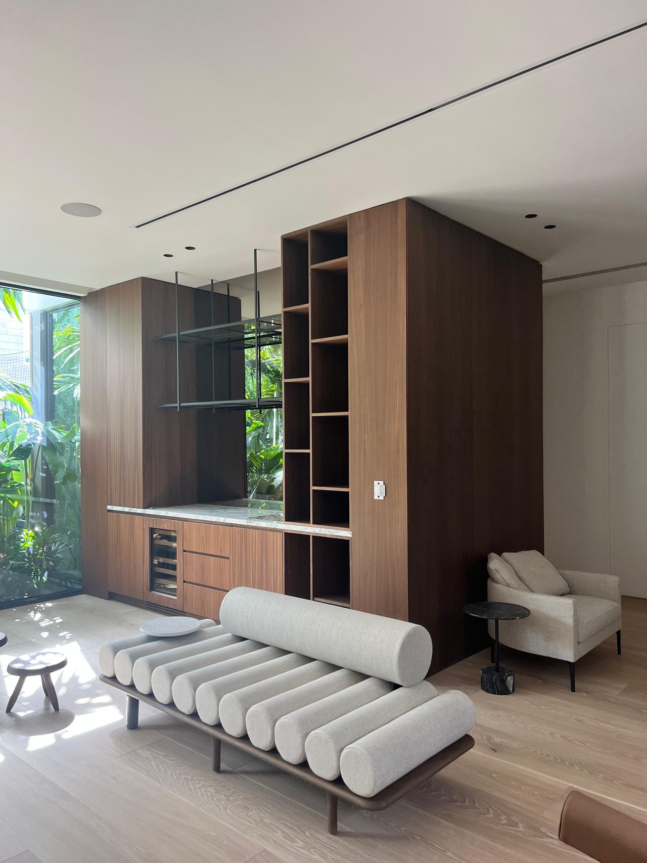 Tall dark wood cabinet tower with open glass shelves, a veined stone countertop section, wine cooler, and adjacent greenery view
