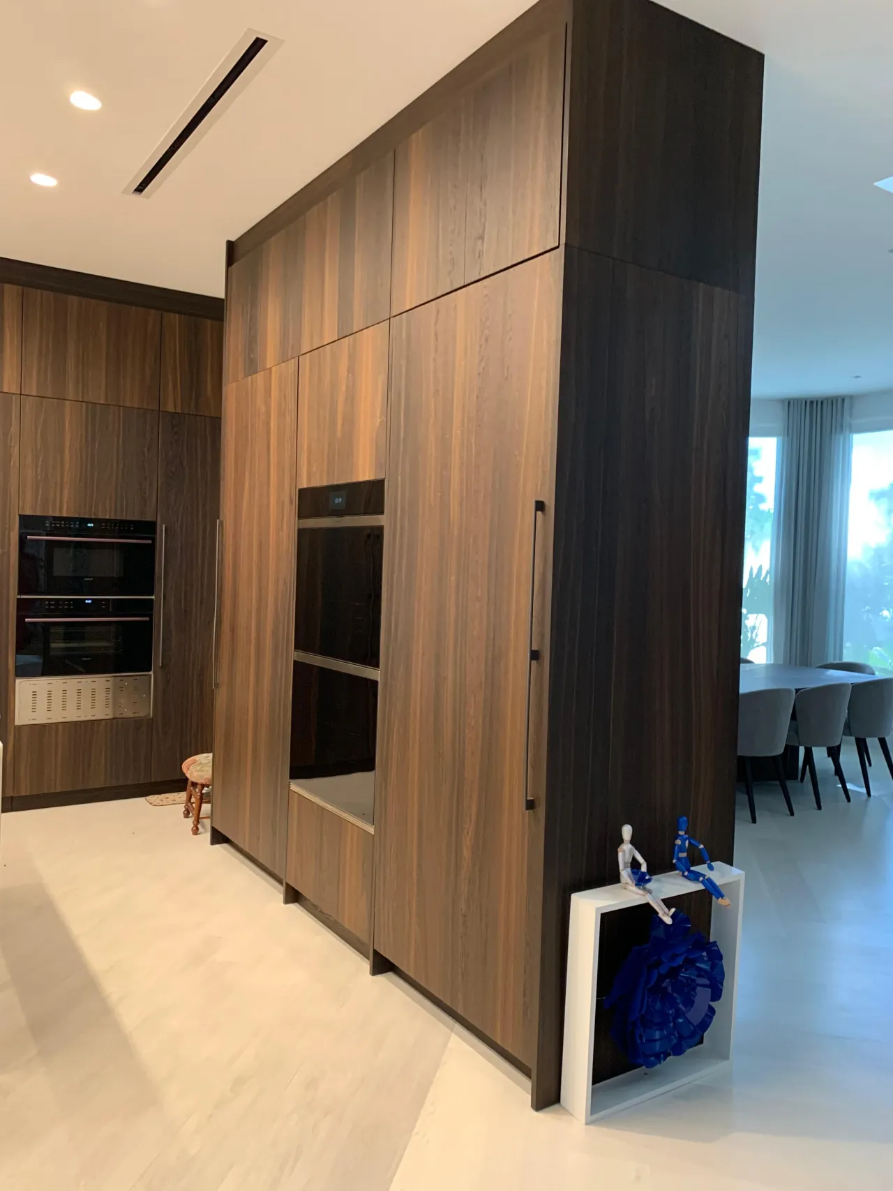 Tall dark wood-grain cabinet tower with built-in wall ovens standing as a room divider, matching cabinetry behind and a dining table beyond