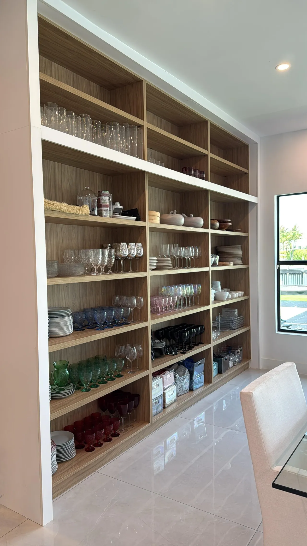 Tall open wood-grain shelving unit with white horizontal dividers, filled with stacked plates and coloured glassware, next to a window