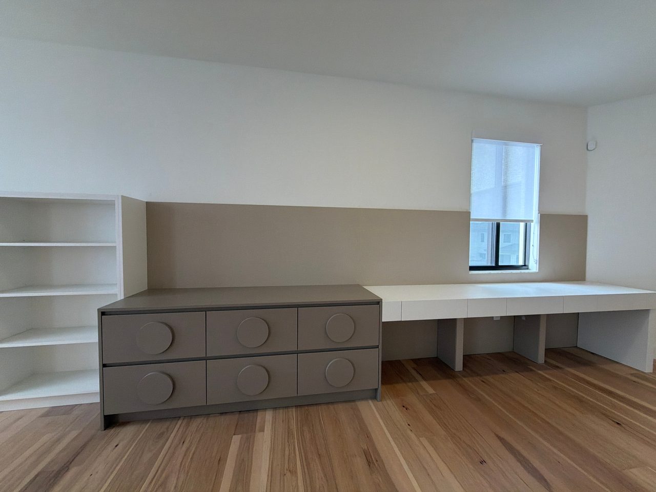 Taupe built-in dresser with circular pulls attached to a floating desk surface, window with roller shade