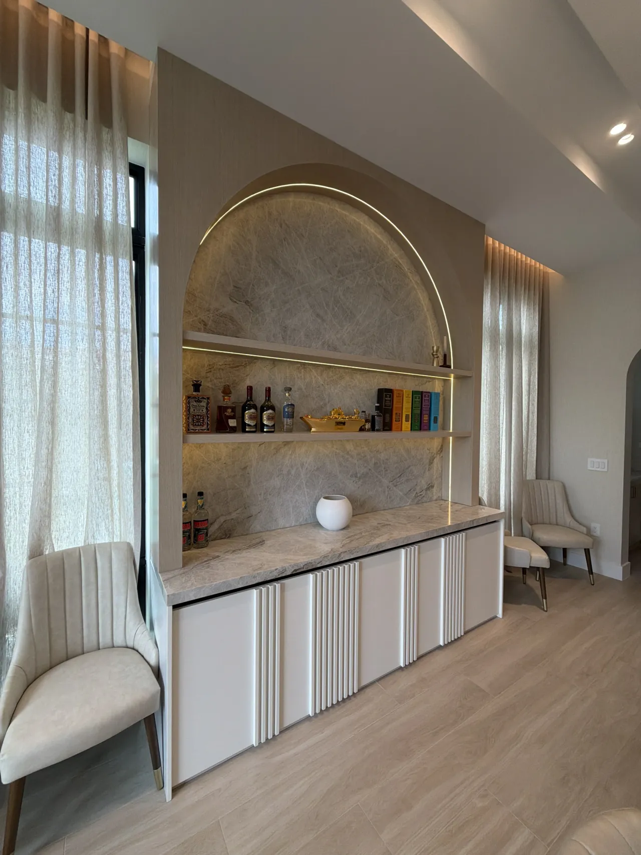 Taupe living room with an arched bar niche lit by gold LED trim over a veined stone backsplash, white fluted credenza below