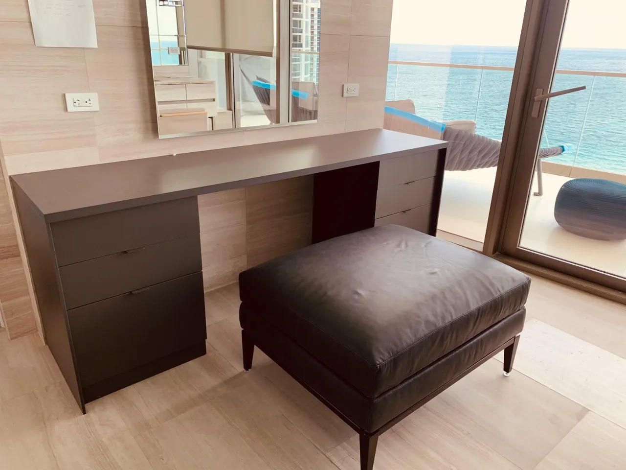 Vanity desk with a dark top and drawer banks flanking an open leg space, dark upholstered ottoman in front, glass door with ocean view at right