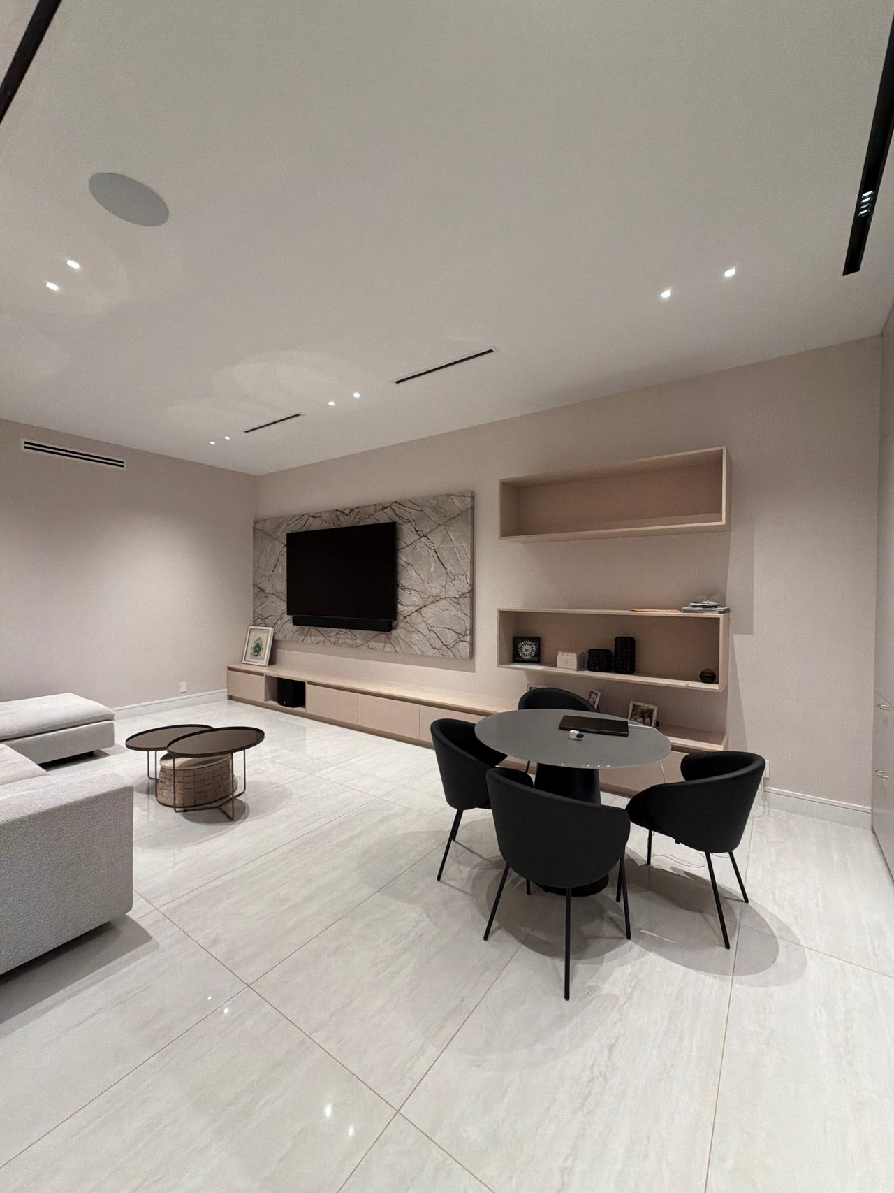 Room with a veined stone wall behind a mounted TV, a long low wood-grain credenza, open wall shelf boxes and a round dark table with black chairs