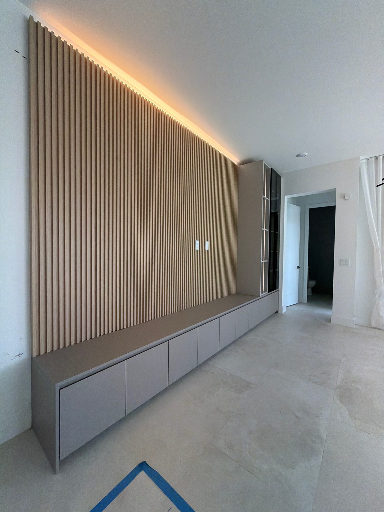 Vertical wood-grain slat feature wall with warm cove lighting above a long low bench with flat-front cabinets, large stone-look floor tiles
