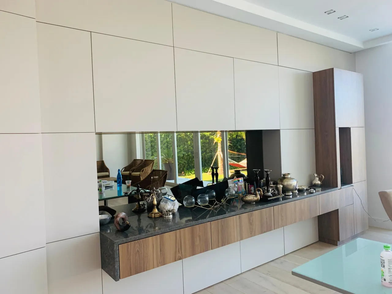 Wall of cream flat-panel cabinetry with a mirrored recess, dark stone counter holding metallic vessels, and wood-grain drawer fronts below