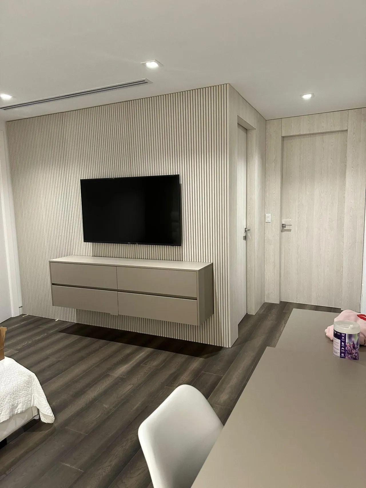 Wall of fine vertical fluted paneling with wall-mounted TV and floating taupe four-drawer dresser, wood-look plank floor and flush wood-grain doors