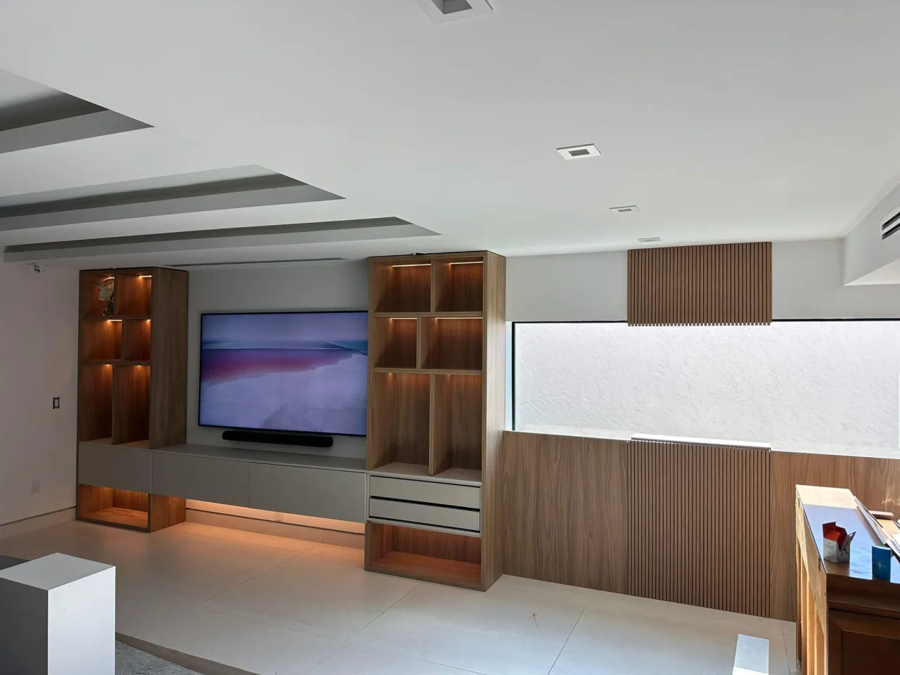 Wall unit with wood-grain shelving towers of lit cubbies flanking a wall-mounted TV on a gray panel, gray media cabinet below with under-shelf lighting