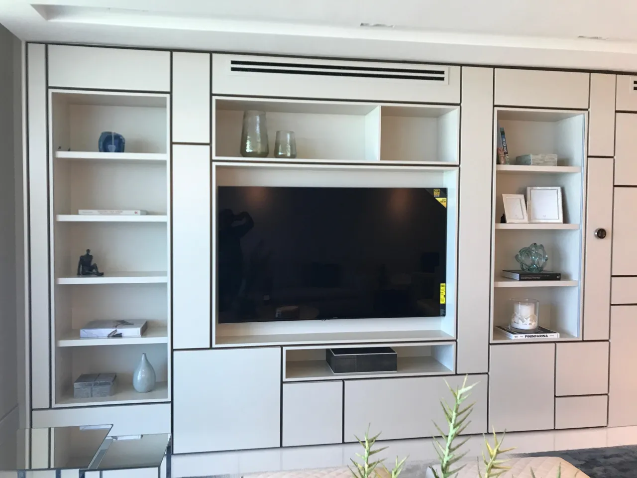 Large white built-in media wall with a recessed mounted TV, open display shelves holding books and objects, and flat-front cabinets and drawers