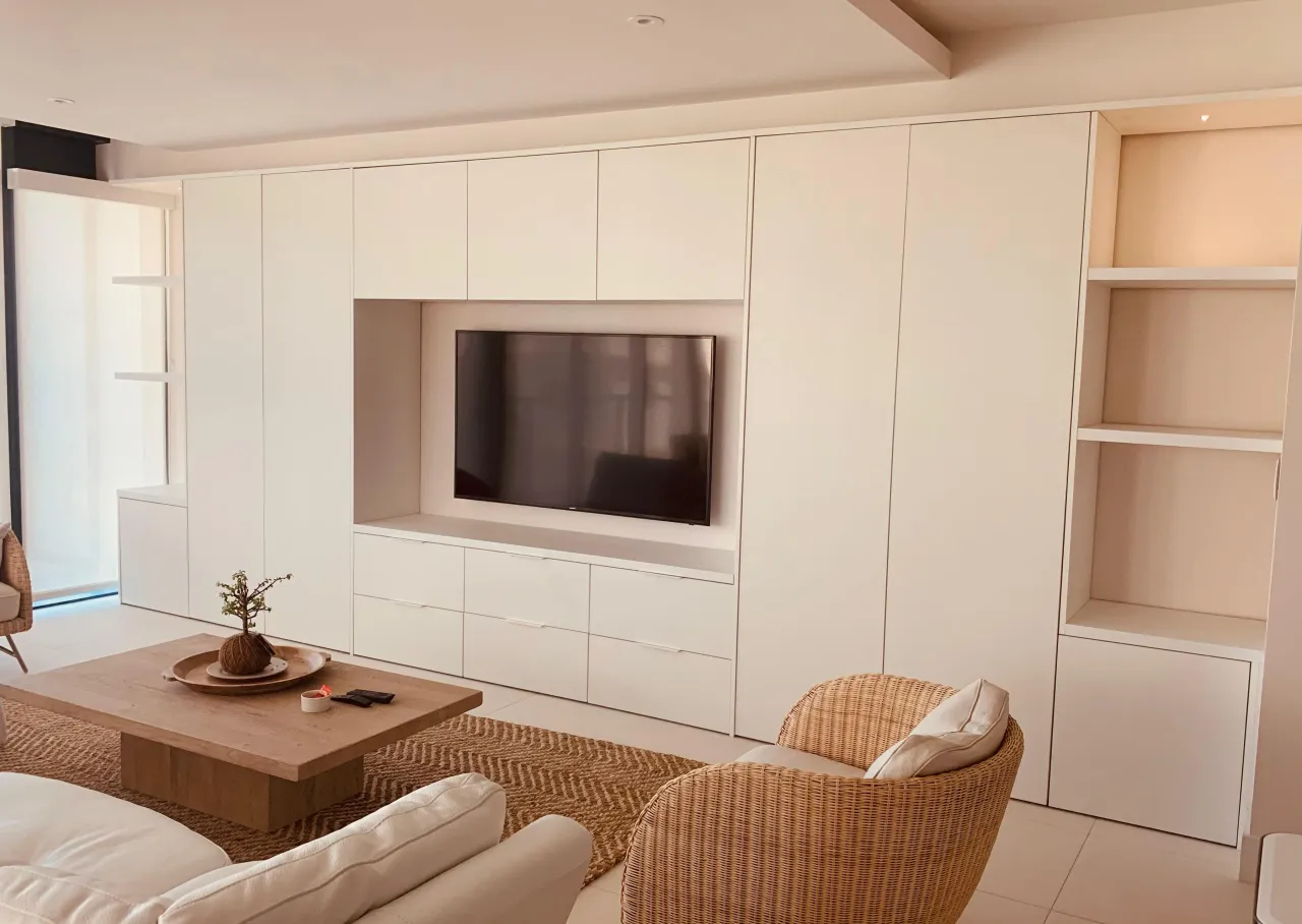 White built-in media wall with a recessed TV niche, tall flat-front doors, drawers and open side shelving, wicker armchairs and a low wood-grain table