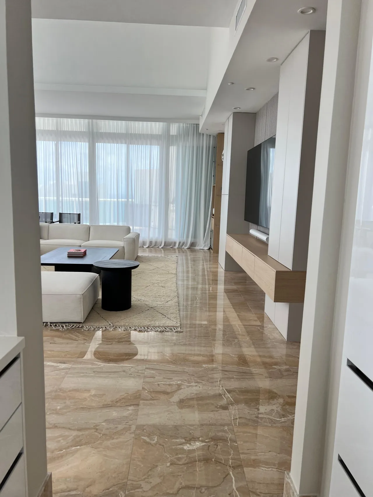 White built-in media wall with wall-mounted TV and light wood-grain floating console, viewed from a hallway across a stone-look tile floor