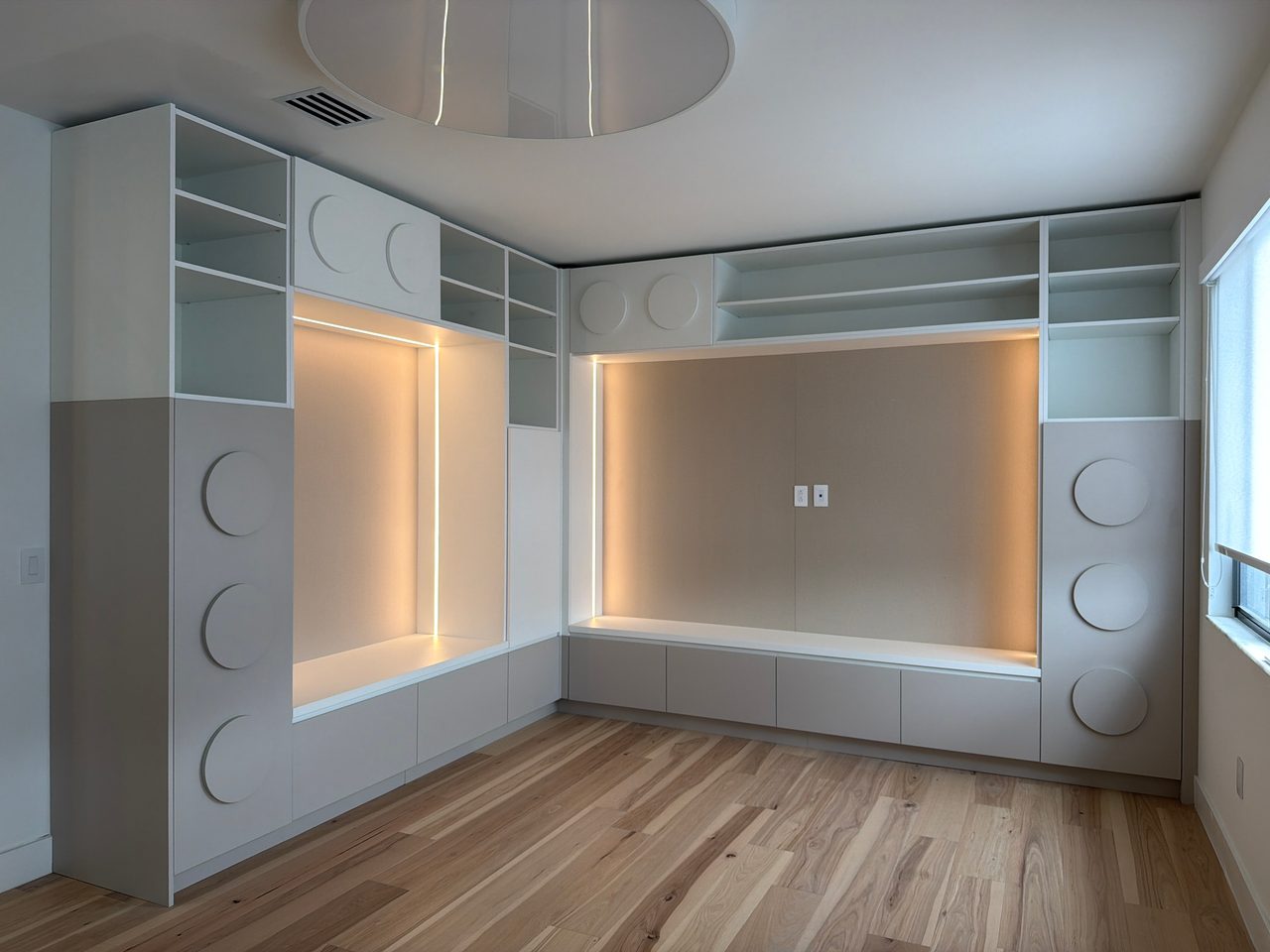 White built-in wall unit wrapping a corner, round disc pulls, open shelving, two backlit niches over a low bench, light wood-grain floor