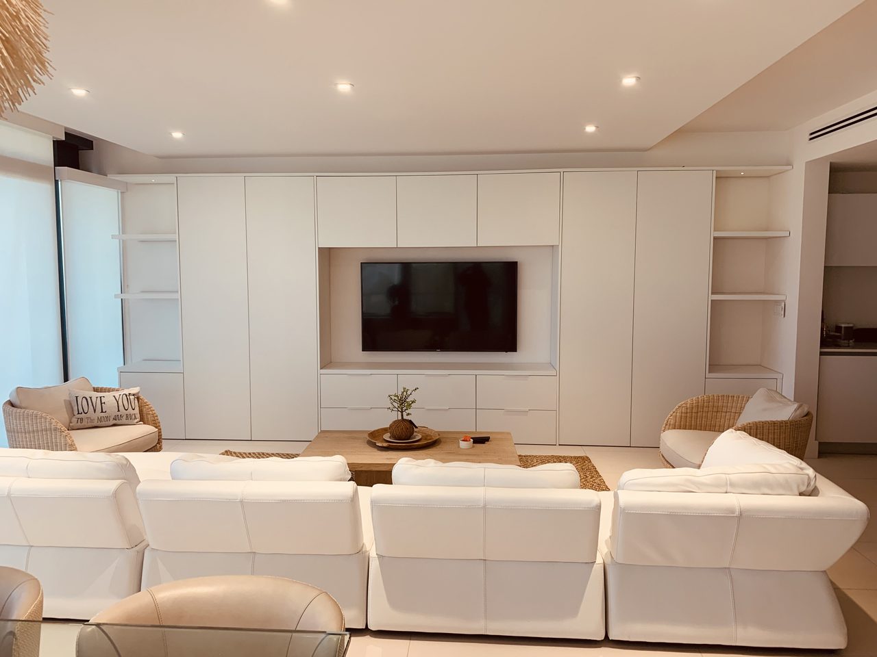 White built-in wall unit with TV niche, drawer bank below and open side shelving, white sofas and woven chairs around a low wood-grain table