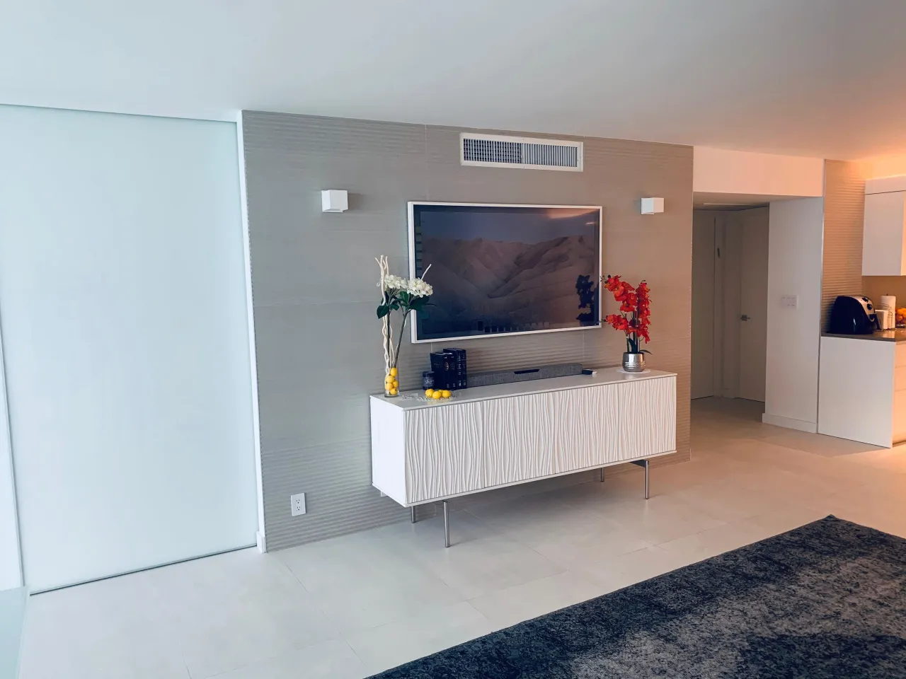 Low white sideboard with rippled textured door fronts on slim metal legs, wall-mounted TV above on a textured taupe wall panel, orchids on top