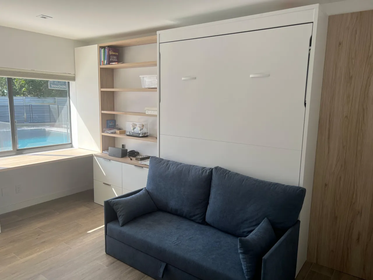 White wall-bed unit with wood-tone open shelving and drawers at left, a blue sofa in front, and a pool visible through the window