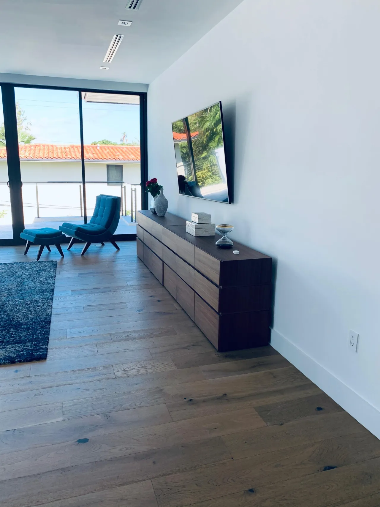 Long low wood-grain drawer credenza against a white wall with a frameless mirror above, plank wood floor, teal lounge chair and ottoman by glass doors