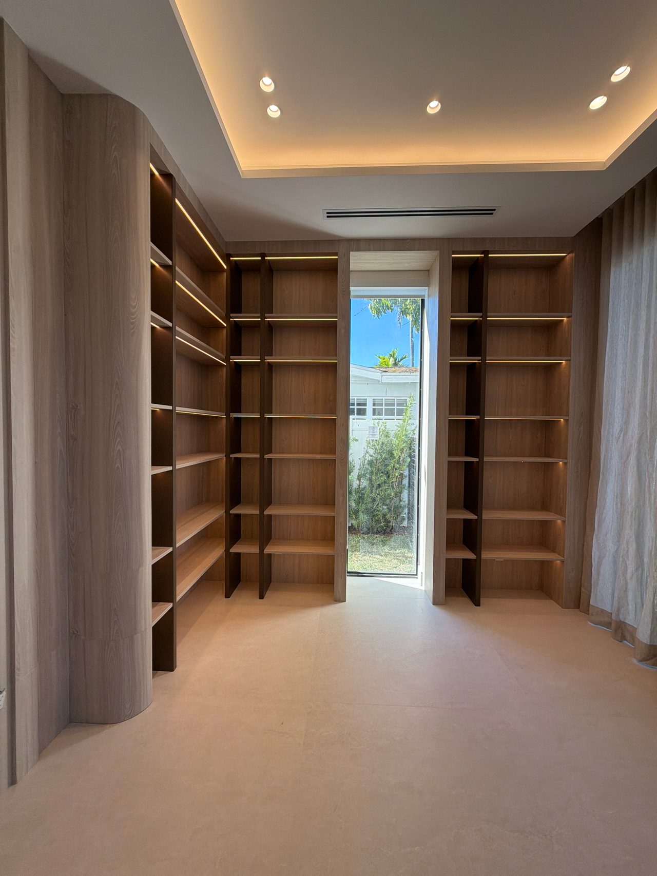 Wood-grain open shelving lining the walls with lit uprights and a cove-lit ceiling, a full-height window showing a palm and a white building