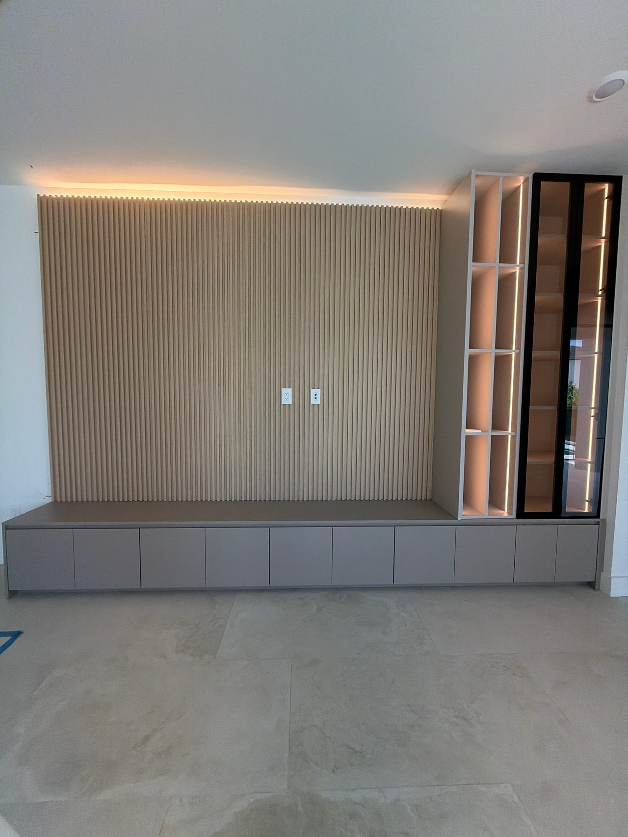 Wood-grain slat wall with cove lighting above a long low cabinet bench, backlit white cubby shelving and a black-framed glass cabinet at right