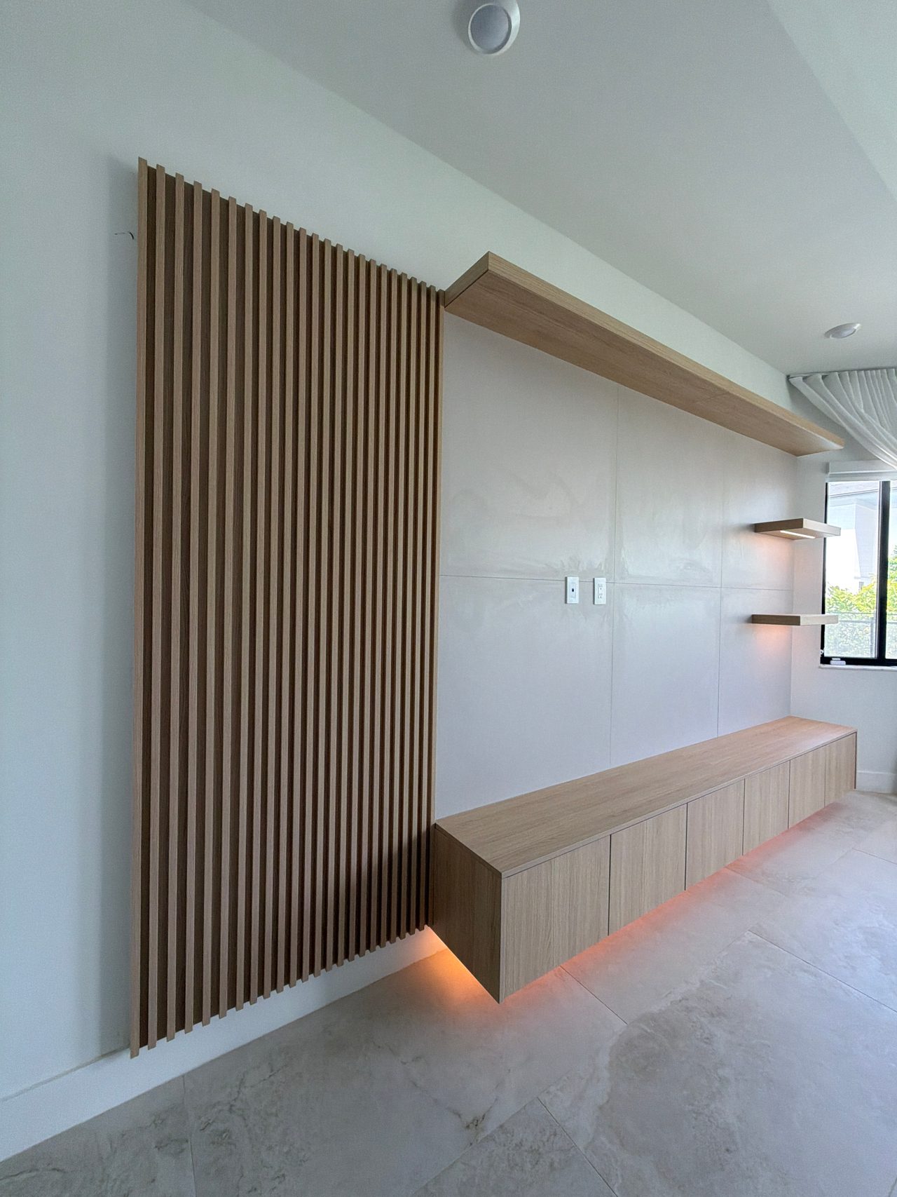 Wood-grain vertical slat wall panel next to a floating wood-grain media cabinet and long upper shelf, with warm LED strip lighting beneath