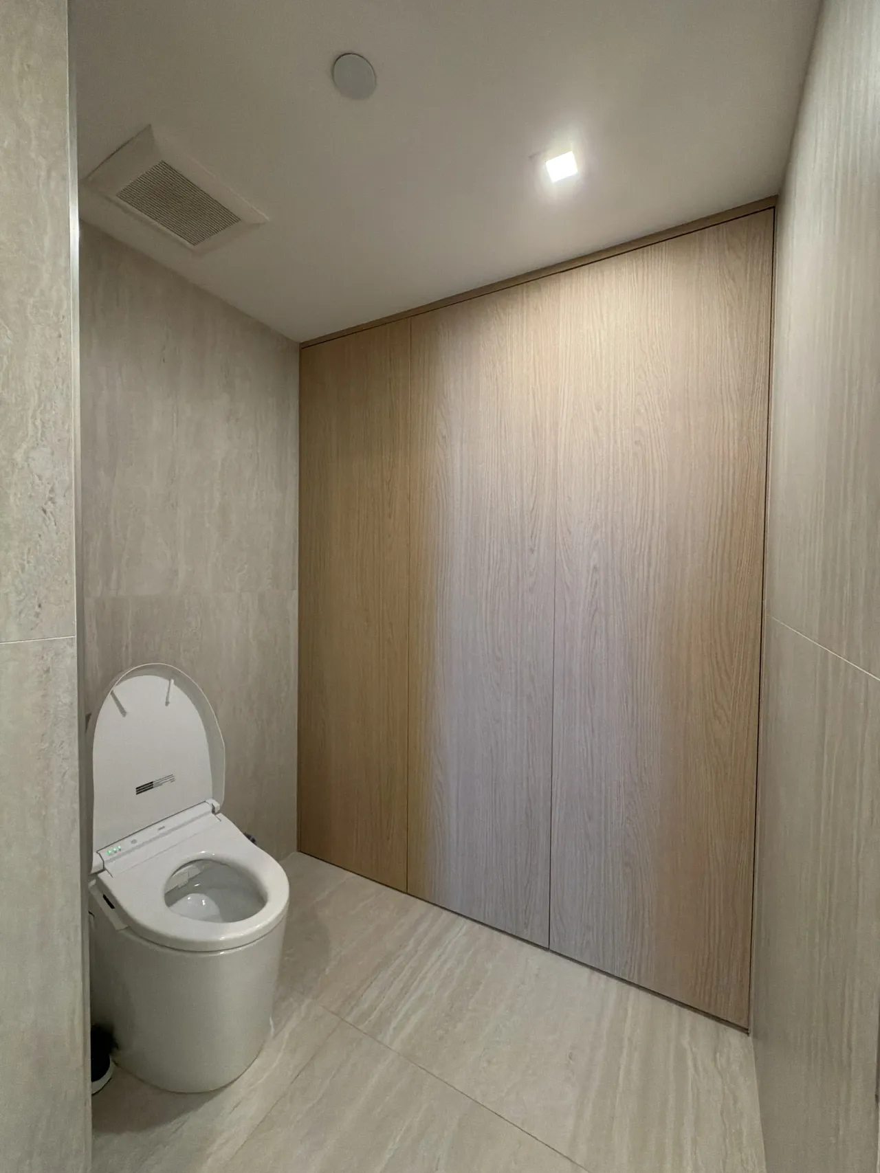 Small bathroom with a toilet beside floor-to-ceiling light wood-grain melamine panel closet doors, light stone tile walls and floor