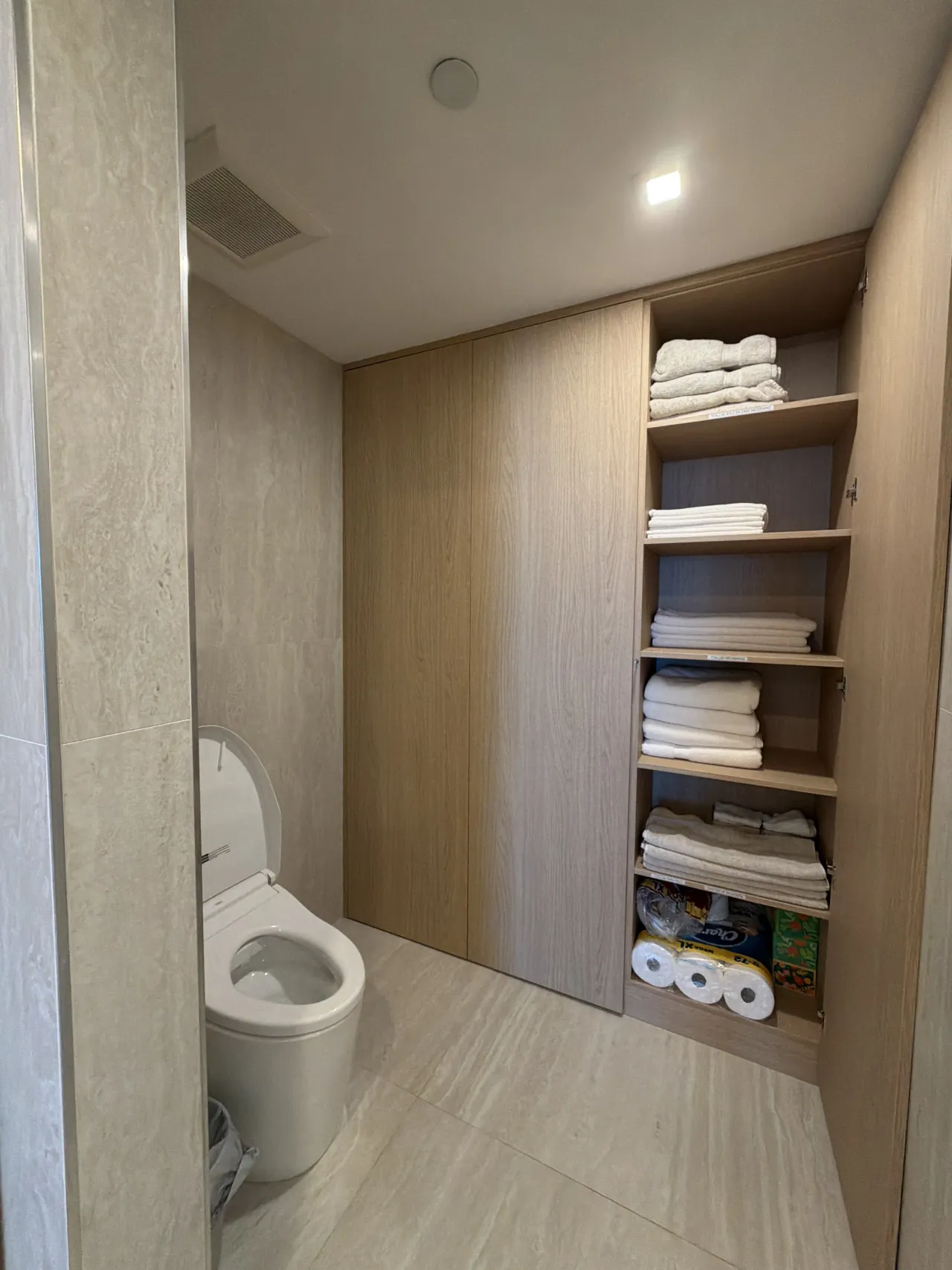 Bathroom with a toilet next to light wood-grain melamine panel closet doors and open linen shelving stocked with folded white towels and paper products
