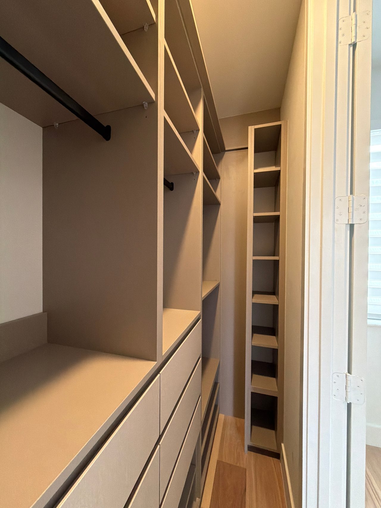Narrow beige walk-in closet with open shelves, a black hanging rod, flat-front drawers and a tall column of small cubbies beside a hinged door
