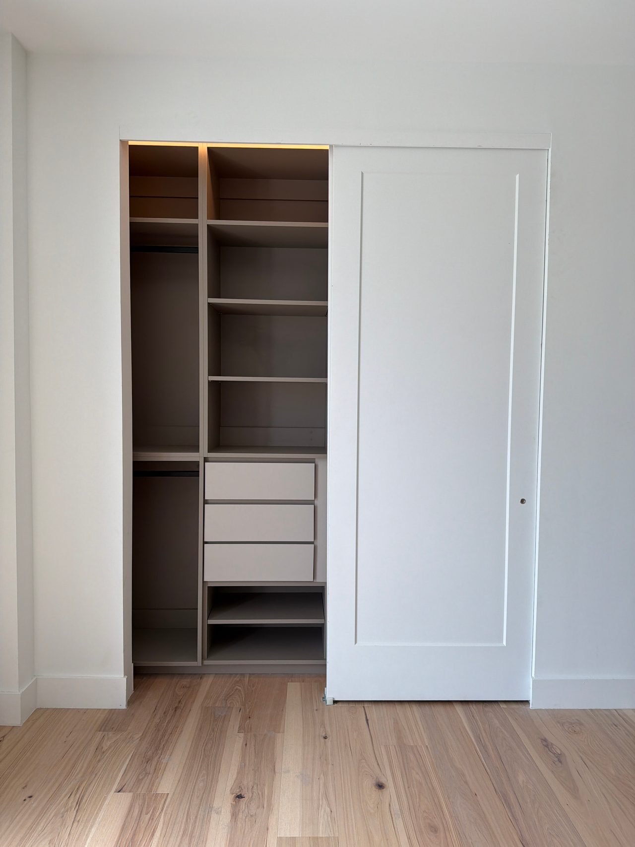 Built-in closet with greige shelves, drawers and hanging rods, half concealed by a white sliding door, over a light wood-grain floor