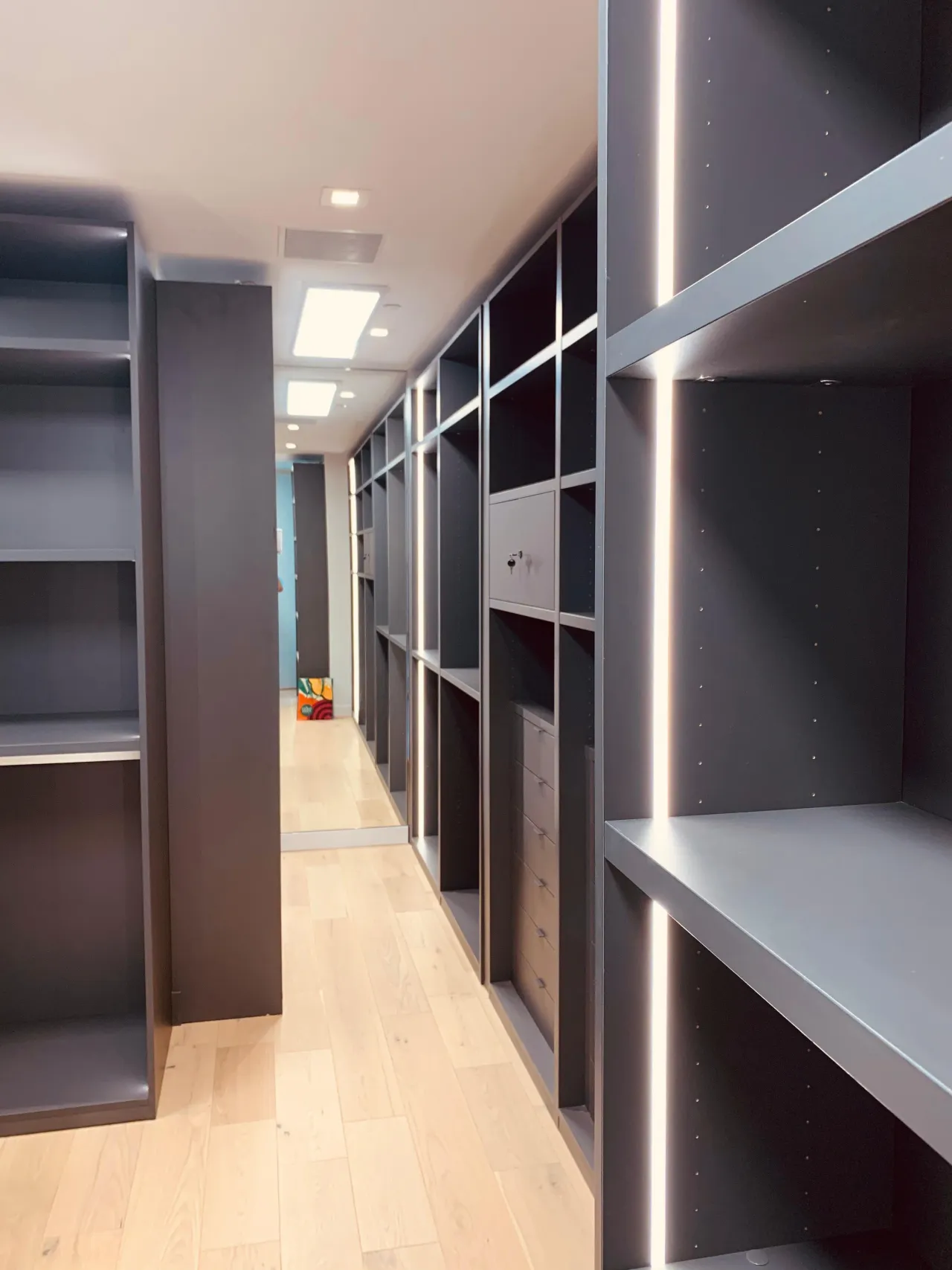 Long charcoal walk-in closet corridor with empty open shelving, LED strip edge lighting, and a small built-in safe visible partway down